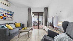 A modern living room with a grey sofa, a coffee table, and a TV. There is a painting on the wall and a view of a dining area through a glass door. - Hola Arnage (Puerto del Carmen)