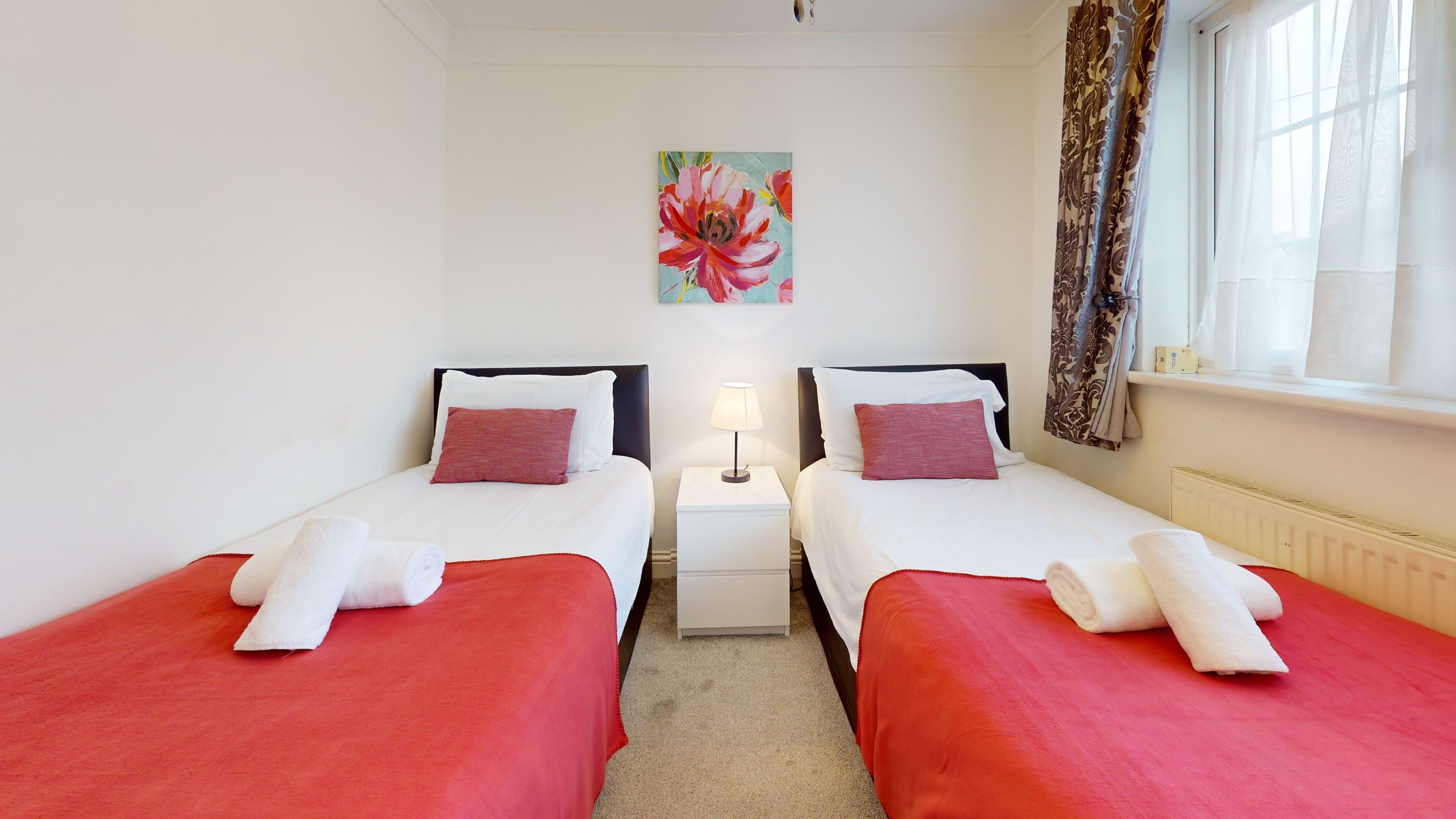 A hotel room with two single beds, each with a red bedspread and white pillows. There is a nightstand with a lamp between the beds, and a painting of a flower on the wall.