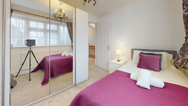 A hotel room with a mirrored wall, a bed with a red bedspread, a nightstand with a lamp, and a chandelier. - Charming 5-bed House in Grays (Grays)