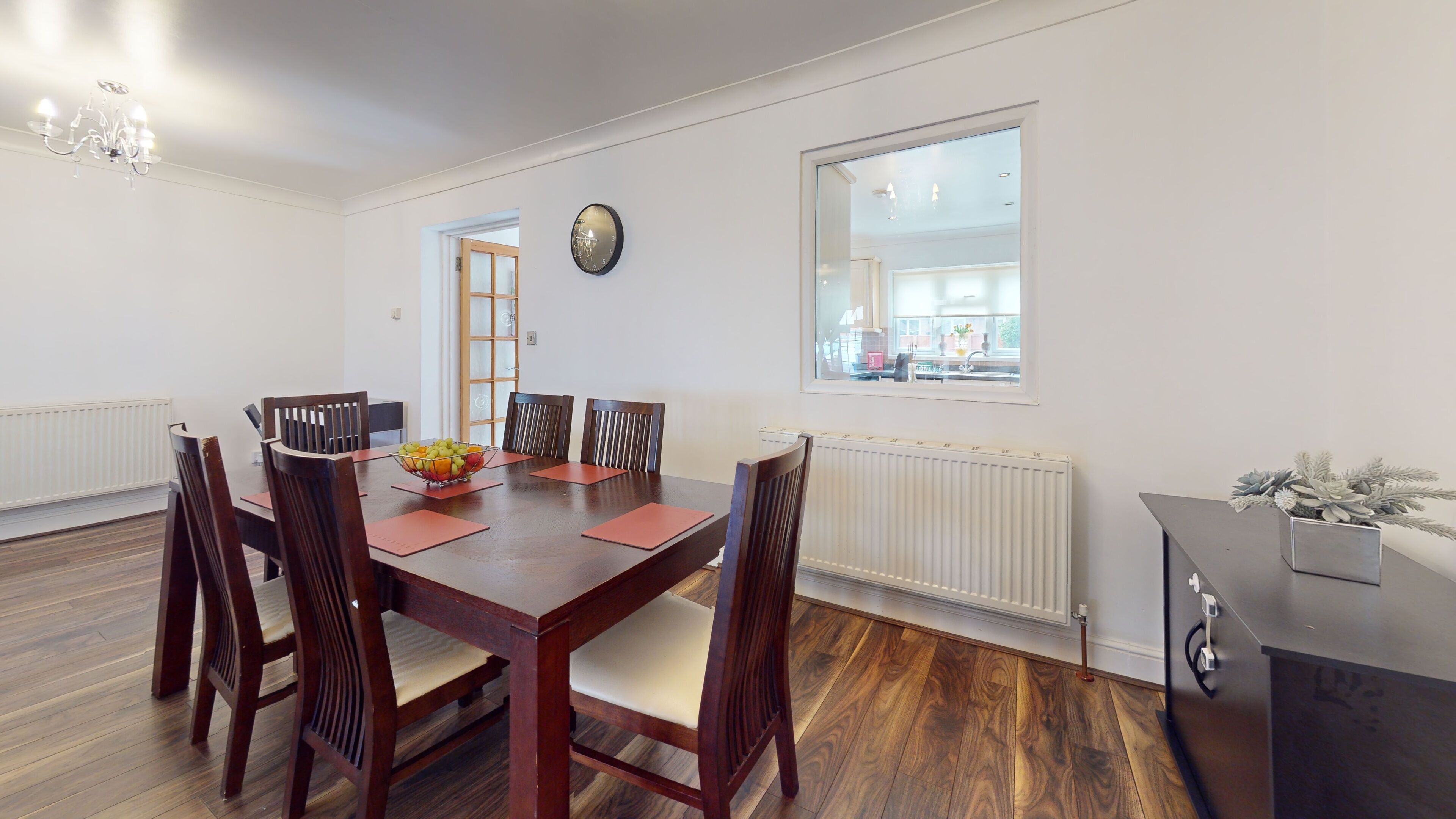 Une salle à manger avec une table en bois, six chaises, un panier de fruits et une vue sur la cuisine par une fenêtre.