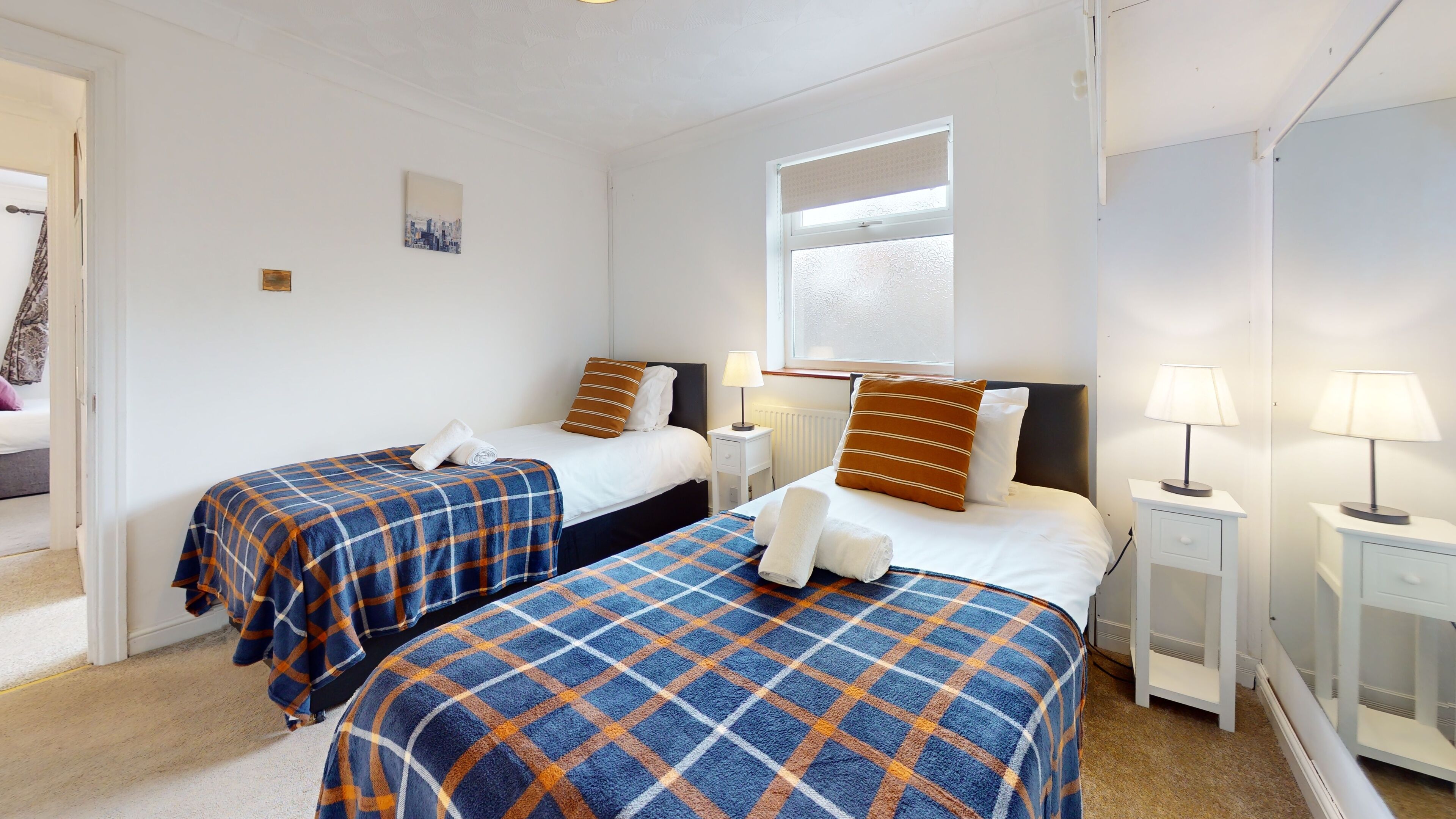 A hotel room with two beds, each with a blue and orange checkered bedspread, white linens, and matching pillows. There are bedside tables with lamps, a window with a view, and a small wall art piece.