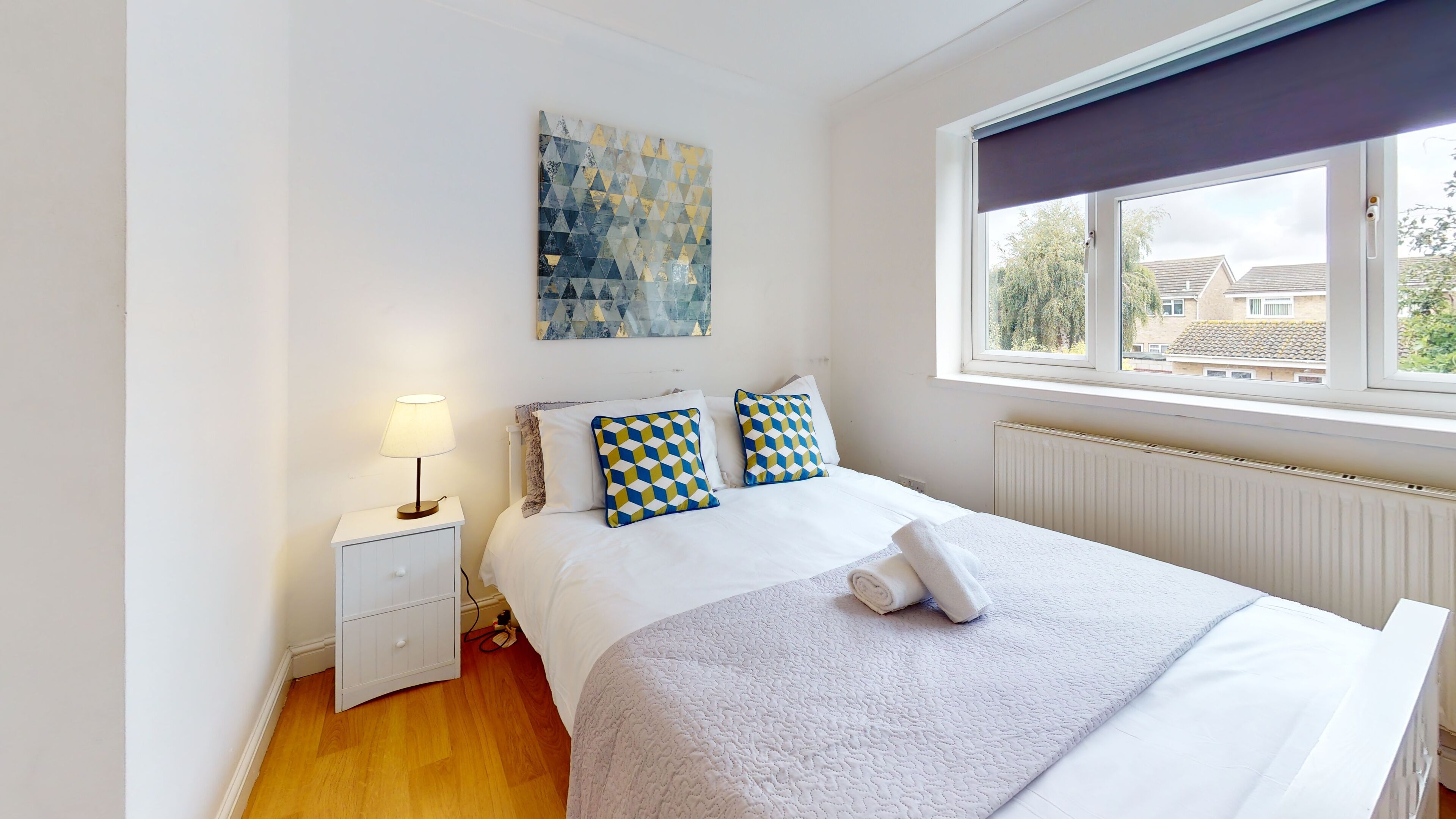 A neatly arranged bedroom with a bed, a bedside table with a lamp, a window with a view of buildings, and a wall with a geometric art piece.