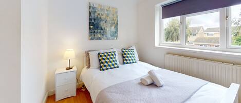 A neatly arranged bedroom with a bed, a bedside table with a lamp, a window with a view of buildings, and a wall with a geometric art piece.