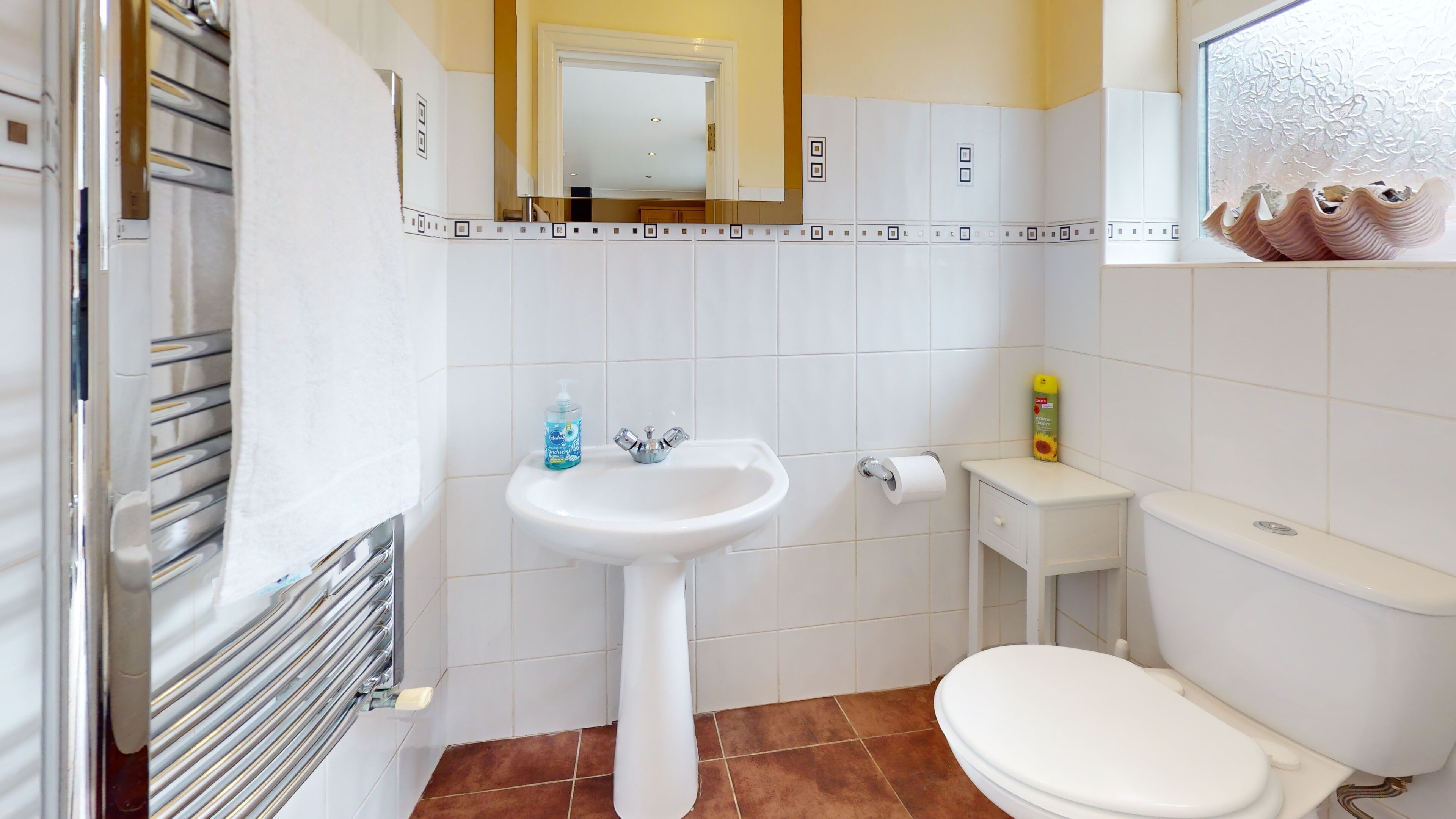 Un baño con un lavabo blanco, inodoro y un espejo sobre el lavabo. Hay un perchero a la izquierda y una pequeña estantería con artículos de aseo.