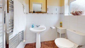 A bathroom with a white sink, toilet, and a mirror above the sink. There is a towel rack on the left and a small shelf with toiletries. - Charming 5-bed House in Grays (Grays)