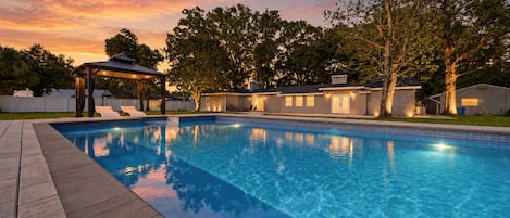 A swimming pool with a covered seating area, surrounded by trees and a building with illuminated windows.