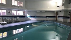 Una piscina interior con agua cristalina, paredes blancas y suelo de baldosas. Hay sillas blancas y una tumbona junto a la piscina.