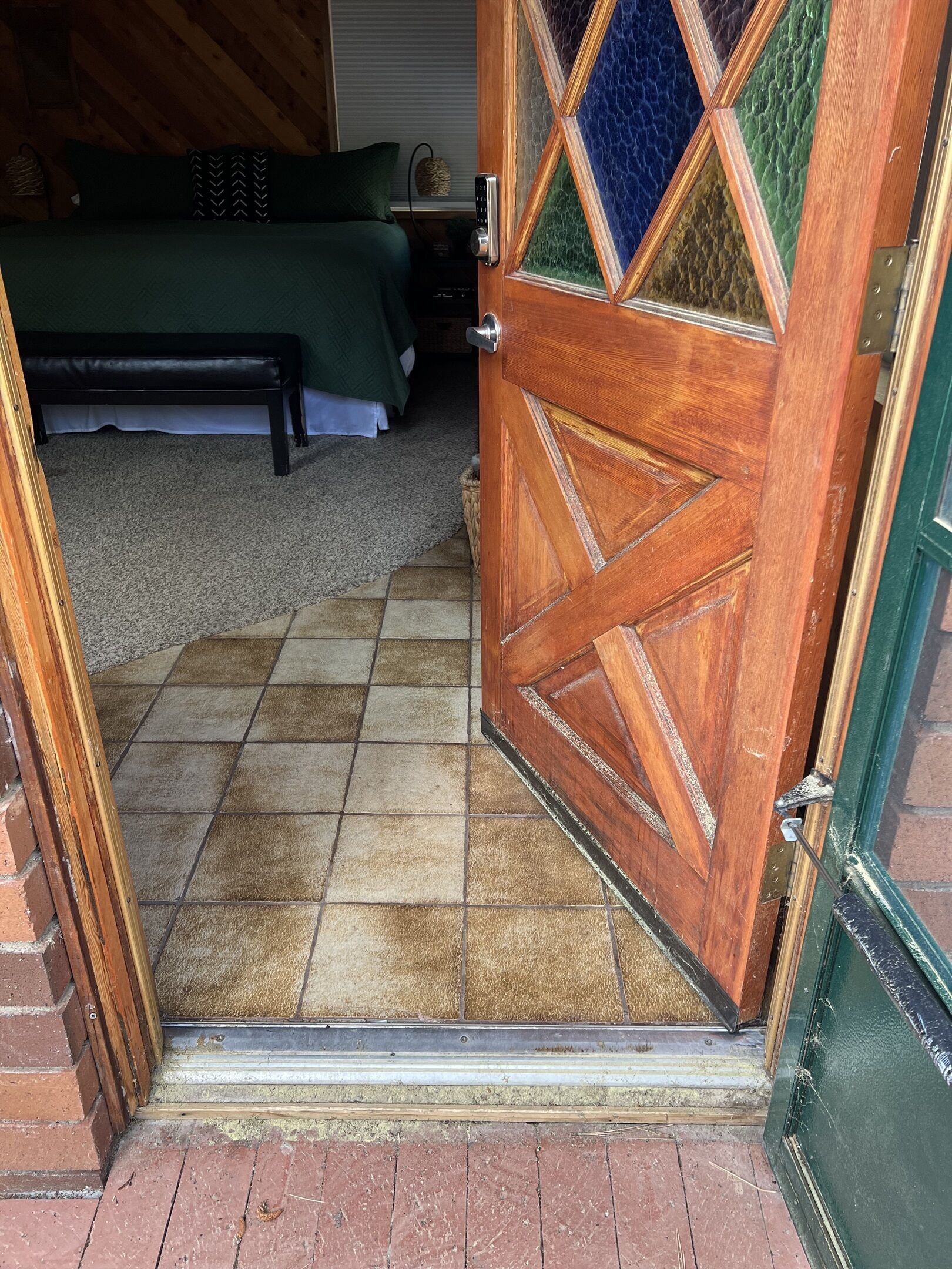 A wooden door with a diamond-patterned glass panel, leading to a room with a green bed and a bench.