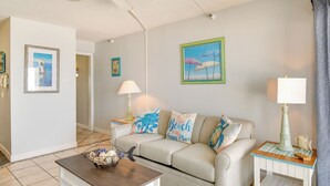 Una sala de estar con un sofá color beige, una mesa de centro, una mesita de noche con una lámpara y decoración en la pared.