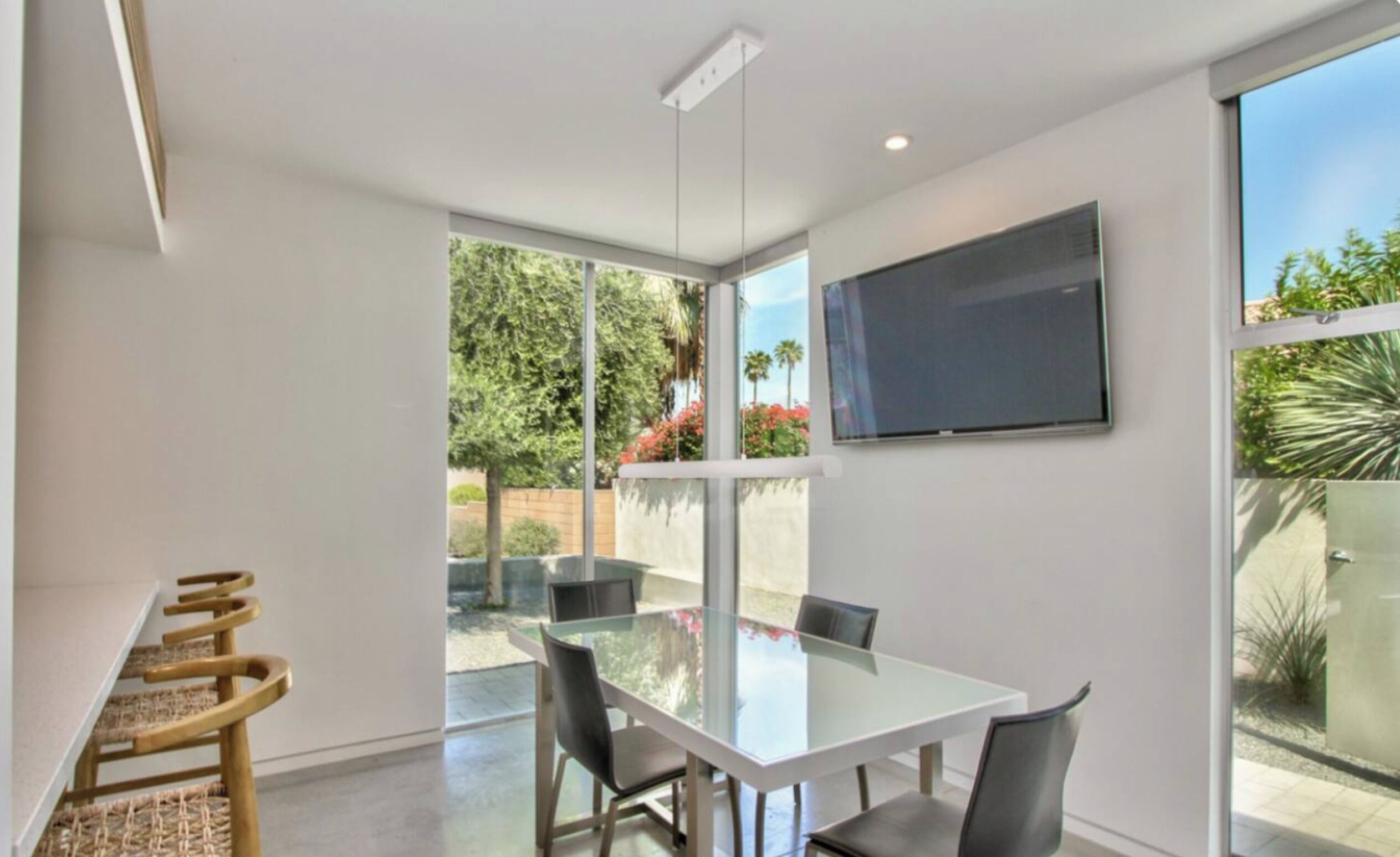 Un comedor moderno con una mesa y sillas de vidrio, un televisor de pantalla plana montado en la pared y grandes ventanas con vistas al exterior.