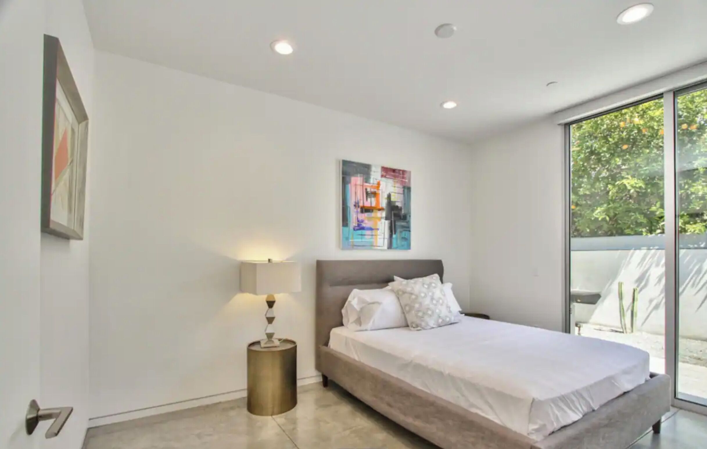 A modern bedroom with a large bed, a bedside lamp, and a window with a view of greenery.