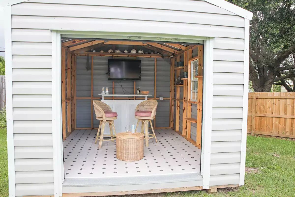 A small, open-sided shed with a television mounted on the wall, two wicker chairs with cushions, and a table with two chairs.
