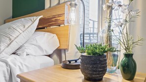 A neatly made bed with white linens, a wooden bedside table with a potted plant, and a clear glass bottle. - Luxury Mid-century Condo, Cozy & Fully Remodeled. Stocked with Many Extras!  (Omaha)