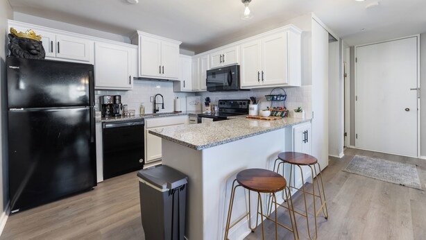 A modern kitchen with white cabinets, a black refrigerator, a microwave, and a granite countertop. There are two stools and a trash can. - Luxury Mid-century Condo, Cozy & Fully Remodeled. Stocked with Many Extras!  (Omaha)