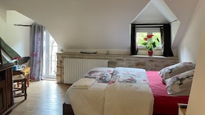 Ein Schlafzimmer mit schrägem Dach, einem Bett mit roter Bettdecke, einem Fenster mit Pflanze und einem Holztisch.