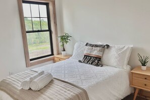 A neatly made bed with a patterned bedspread, a bedside plant, and a window with a view of greenery.