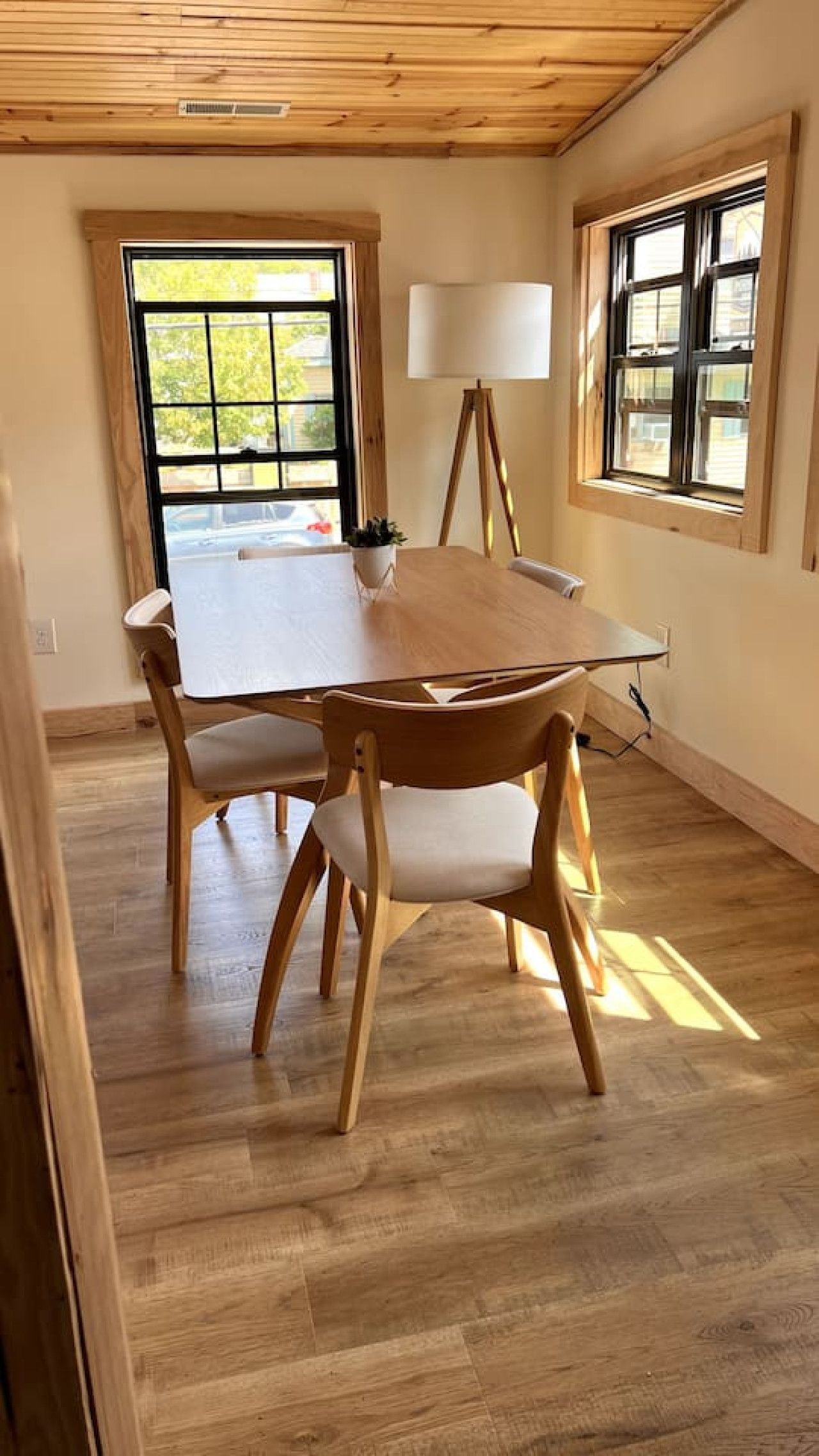 Un comedor con mesa y sillas de madera, una lámpara de pie y ventanas amplias con vista a la vegetación exterior.