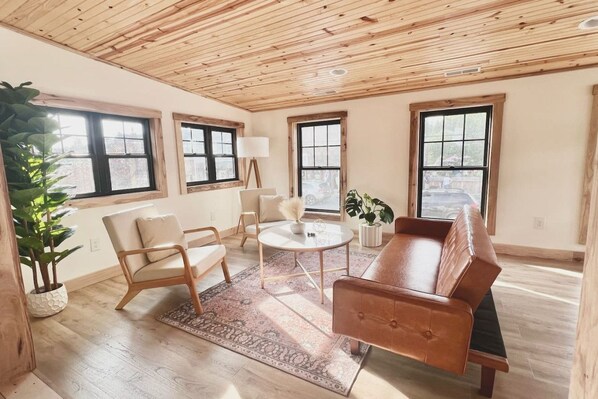 A spacious living room with a wooden ceiling, large windows, a sofa, a coffee table, and a floor lamp.