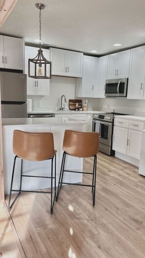 Una cocina moderna con armarios blancos, un refrigerador de acero inoxidable, un microondas y un horno doble. Hay dos taburetes marrones en la isla de la cocina.