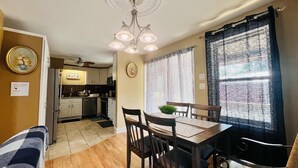 Una cocina compacta con armarios blancos, un microondas y un lavamanos. Hay una zona de comedor con una mesa y sillas, y una planta pequeña sobre la mesa.
