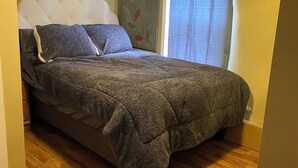 Un dormitorio con cama, piso de madera, cabecera y ventana con cortinas.