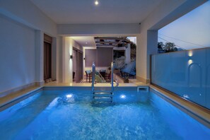 Une piscine avec un mur en verre, une terrasse ensoleillée avec des sièges d’extérieur et une vue sur une colline.