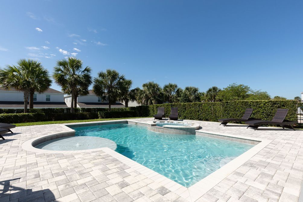 A rectangular swimming pool with a hot tub, surrounded by lounge chairs and palm trees.