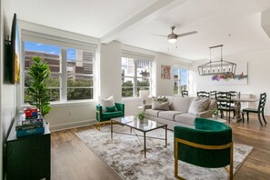 Amplia sala de estar con sofá, mesa de centro y sillas, ventanas grandes y un comedor con candelabro.