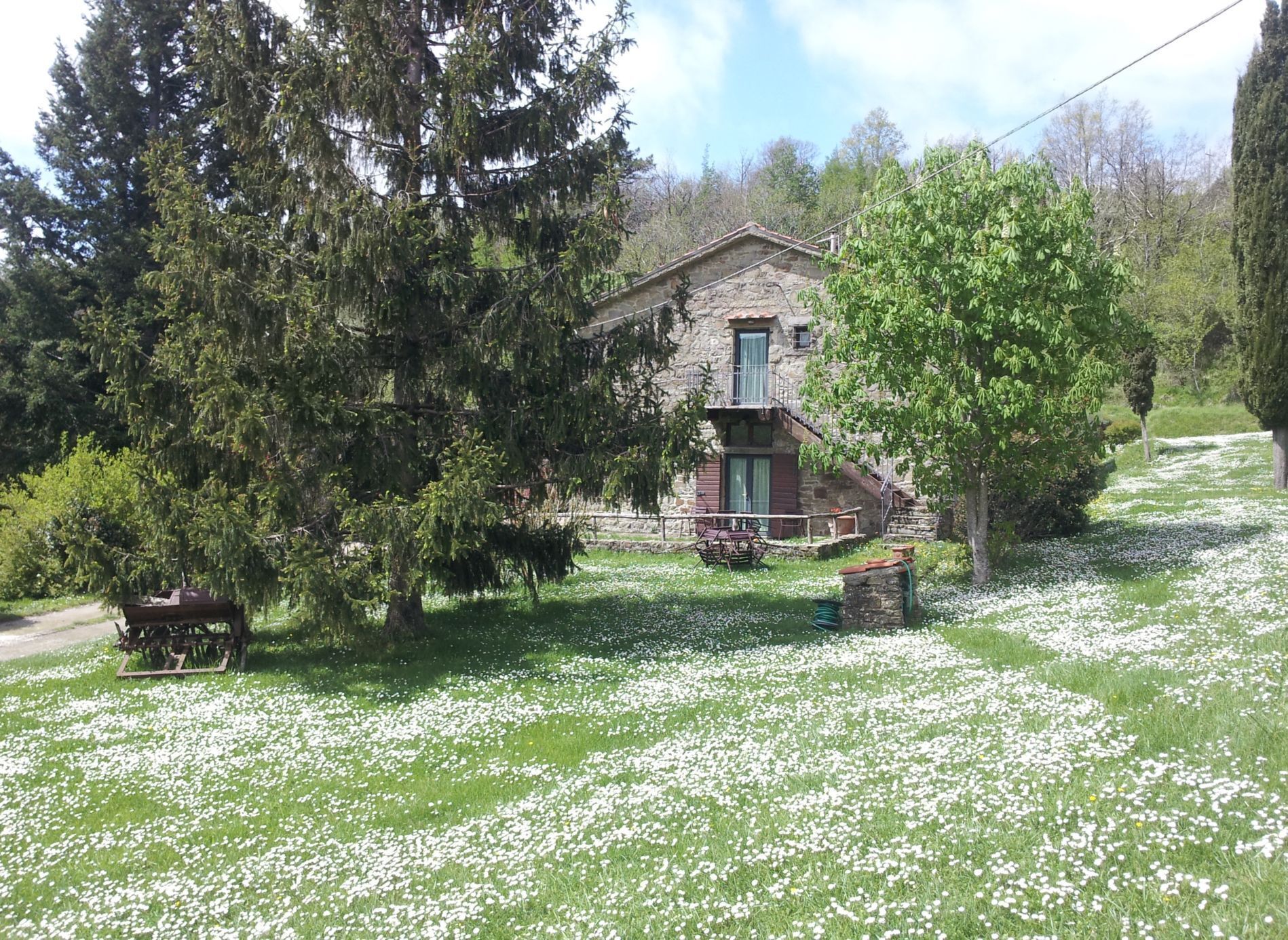 A stone building with a balcony, surrounded by trees and a grassy area with white flowers.