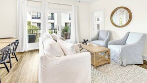Un salon spacieux avec un canapé blanc, un fauteuil gris, une table basse en bois et une grande fenêtre donnant sur un balcon.