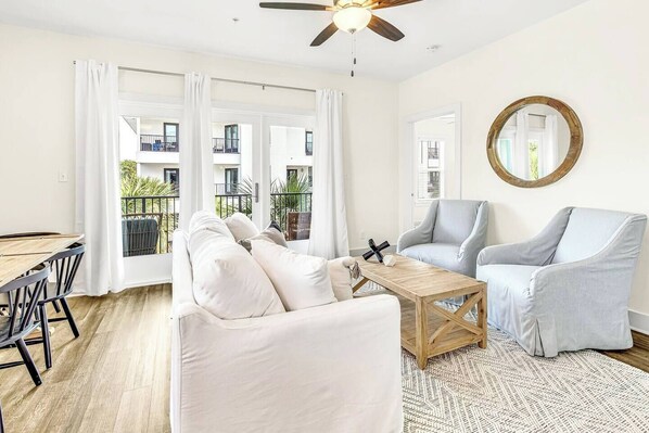 A spacious living room with a white sofa, a grey armchair, a wooden coffee table, and a large window with a view of a balcony.