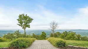 A paved pathway leading through a landscaped area with trees and shrubs, overlooking a distant landscape. - Private 12-Acre Mountain Estate with Blue Ridge Views, Game Room & Sleeps 20 (Fletcher)