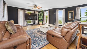 A living room with leather sofas, a TV, a ceiling fan, and a large window with a view. - Private 12-Acre Mountain Estate with Blue Ridge Views, Game Room & Sleeps 20 (Fletcher)