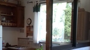 A kitchen with wooden cabinets, a sink, and a window with a view of greenery. - Charmante Maisonnette au Calme (Montmeyan)