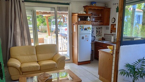 A living room with a sofa, a wooden coffee table, and a kitchen area with wooden cabinets and a refrigerator. - Charmante Maisonnette au Calme (Montmeyan)