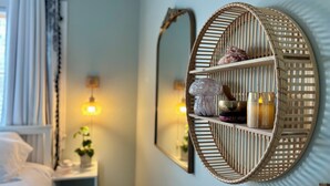 A room with a circular woven shelf holding decorative items, a mirror, and a bed with white bedding.