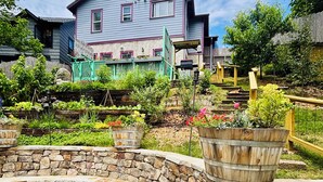 A stone garden with a curved stone wall, a wooden barrel, and a house with a blue roof and pink trim in the background.