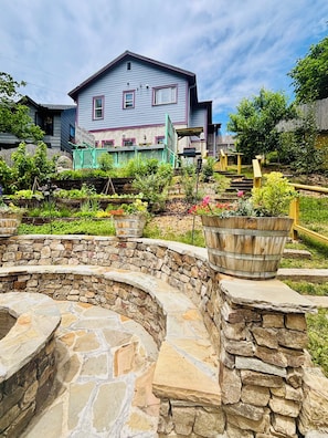 Ein Steingarten mit einer gebogenen Steinmauer, einem Holzfass und einem Haus mit blauem Dach und rosa Verzierungen im Hintergrund.