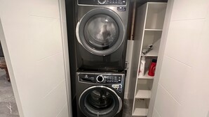 Une petite buanderie équipée de deux machines à laver à chargement frontal superposées, d’une étagère contenant des produits de nettoyage et d’un sol carrelé en marbre.