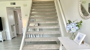 Un escalier avec des marches recouvertes de moquette, une console blanche surmontée d’un vase de fleurs et un tableau encadré.