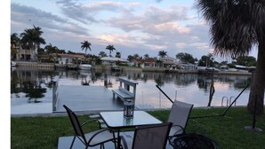 Un patio al aire libre con mesa y sillas, un hogar de fuego metálico y una vista de un lago tranquilo con barcos y casas al fondo.