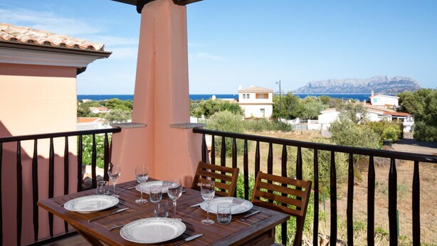 Un balcon avec une table dressée pour deux personnes, d’où l’on peut admirer une ville côtière avec ses maisons et une montagne en arrière-plan.