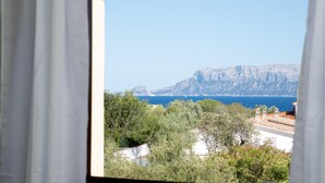 Une vue depuis une fenêtre avec des rideaux, donnant sur un paysage côtier avec des montagnes et une mer bleue et limpide.