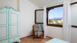 Une chambre avec un lit, une chaise, une armoire et une fenêtre donnant sur la mer et les montagnes.
