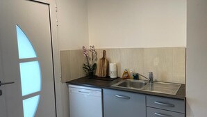 A compact kitchen with a dishwasher, a sink, and a countertop with a coffee maker, a bottle of dish soap, and a vase with flowers. - Chambre Giverny (Hargeville)