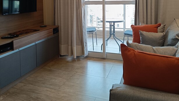 Ett modernt vardagsrum med en platt-TV, träinredning, en soffa med orange kuddar och en skjutdörr i glas som leder ut till ett utomhusområde.