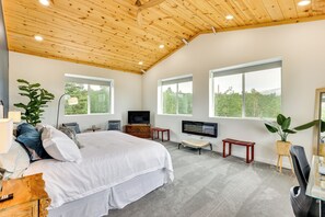 Un dormitorio espacioso con una cama grande, televisión, techo de madera y ventanas amplias con vistas a zonas verdes.
