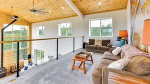 A spacious living area with a high ceiling, wooden beams, a balcony with a railing, and a large window offering a view of the outdoors. - Colorado Retreat w/ Gas Grill & Mountain Views! (Fairplay)