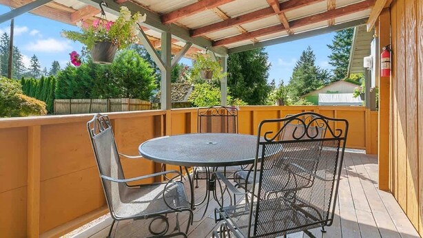 Un patio techado con una mesa redonda de metal y dos sillas, rodeado de barandillas de madera y plantas en macetas.