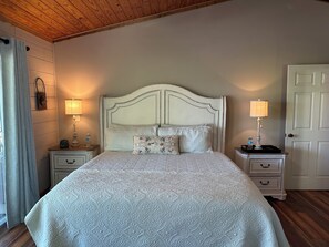 Un dormitorio con una cama grande, dos mesitas de noche, techo de madera, una puerta blanca y una ventana con cortinas.